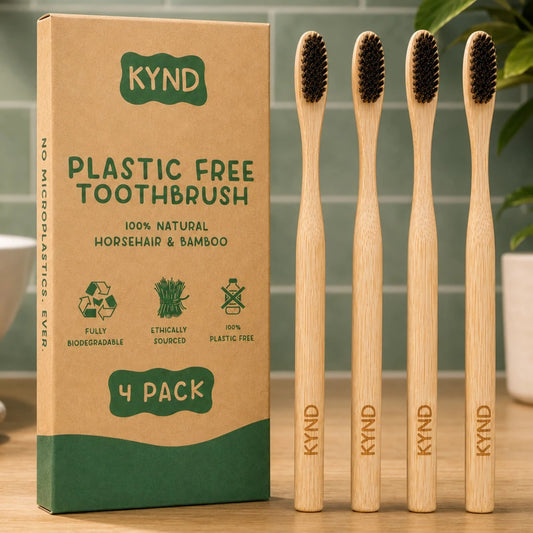Four KYND plastic-free bamboo toothbrushes with dark bristles stand next to their brown kraft paper packaging box on a wooden surface.