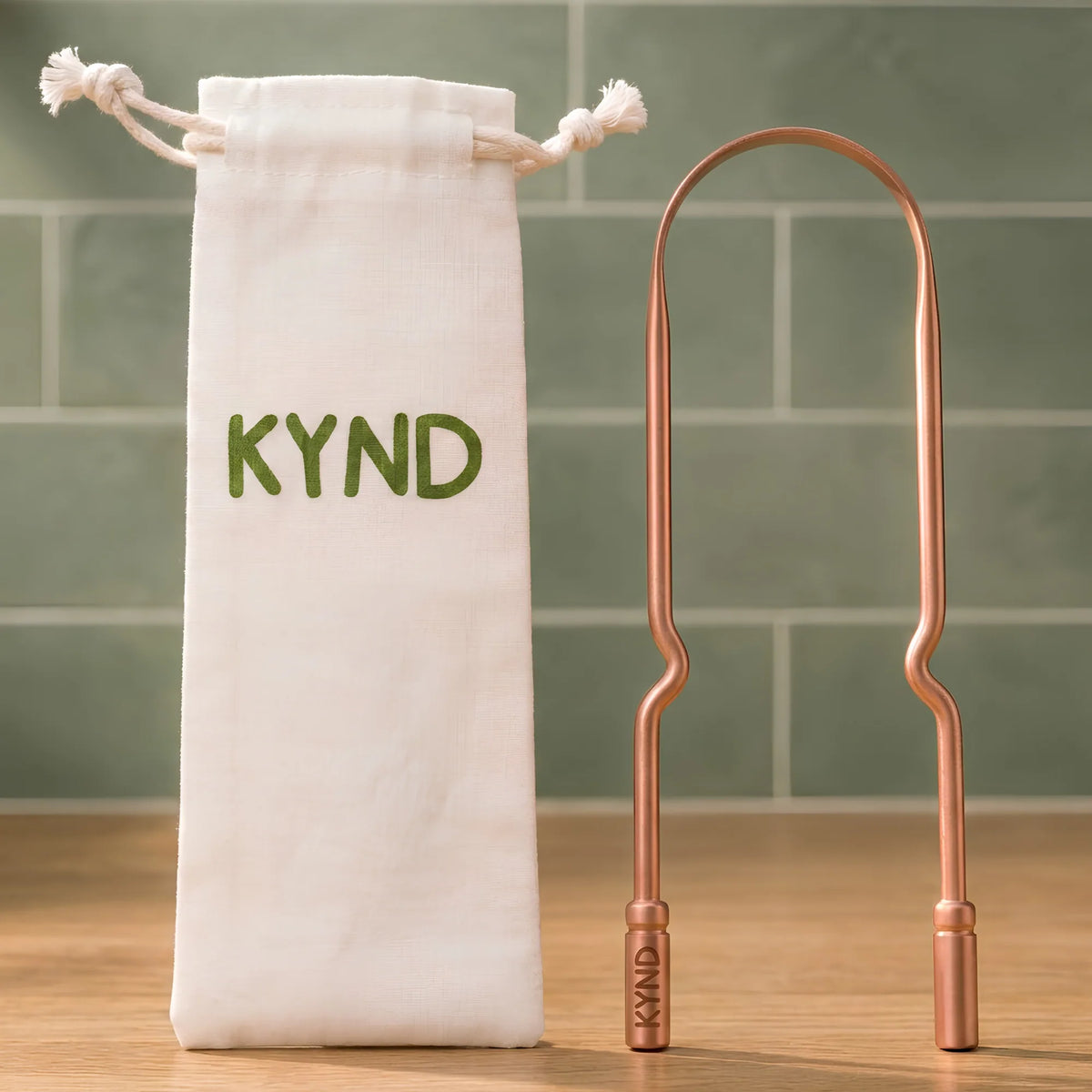 A copper-colored KYND personal tongue scraper and its matching white drawstring pouch with green KYND lettering, set against a green tiled background.