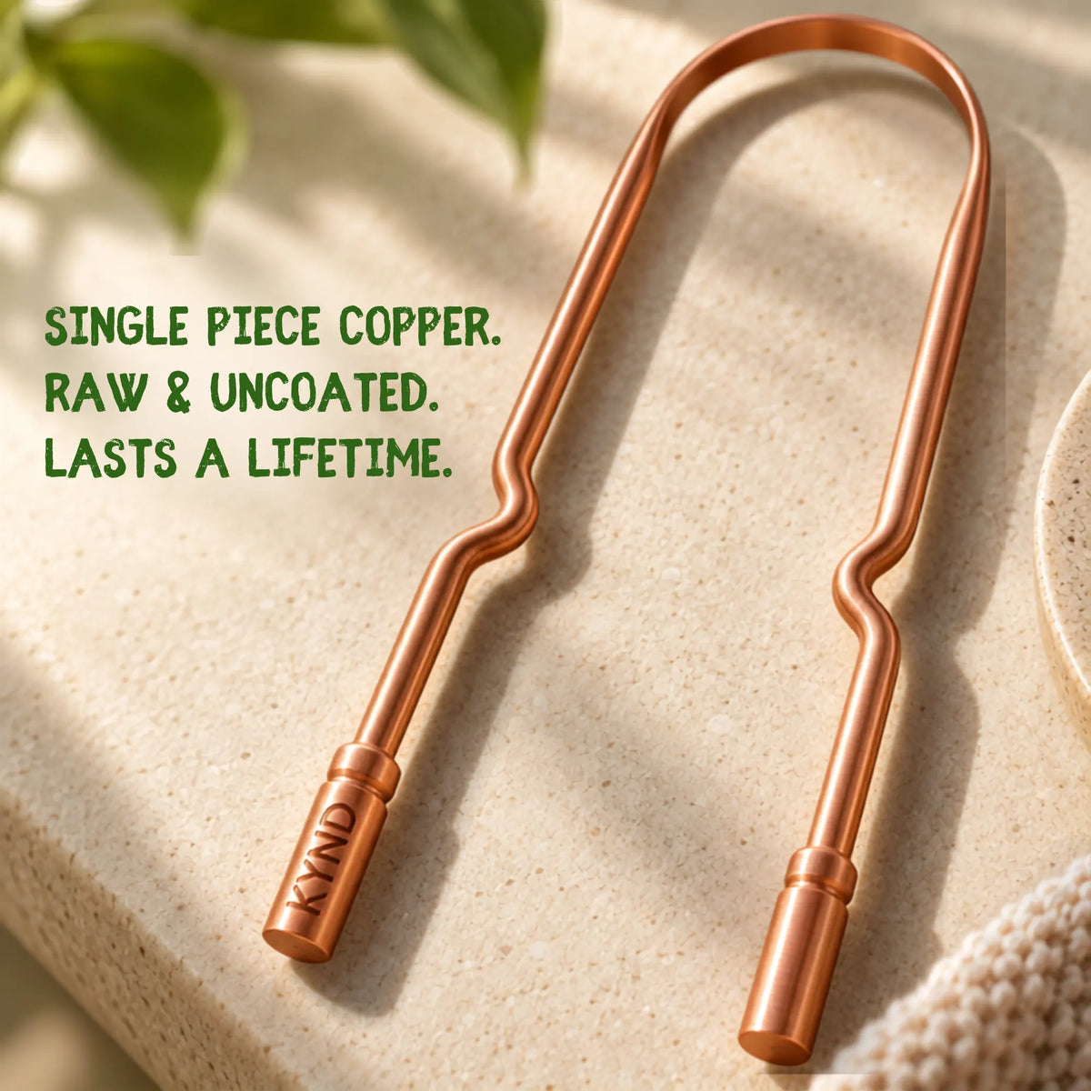 A copper tongue scraper with a wavy design, shown on a textured beige stone surface with green leaves in the background. Text on the image reads 'SINGLE PIECE COPPER. RAW & UNCOATED. LASTS A LIFETIME.