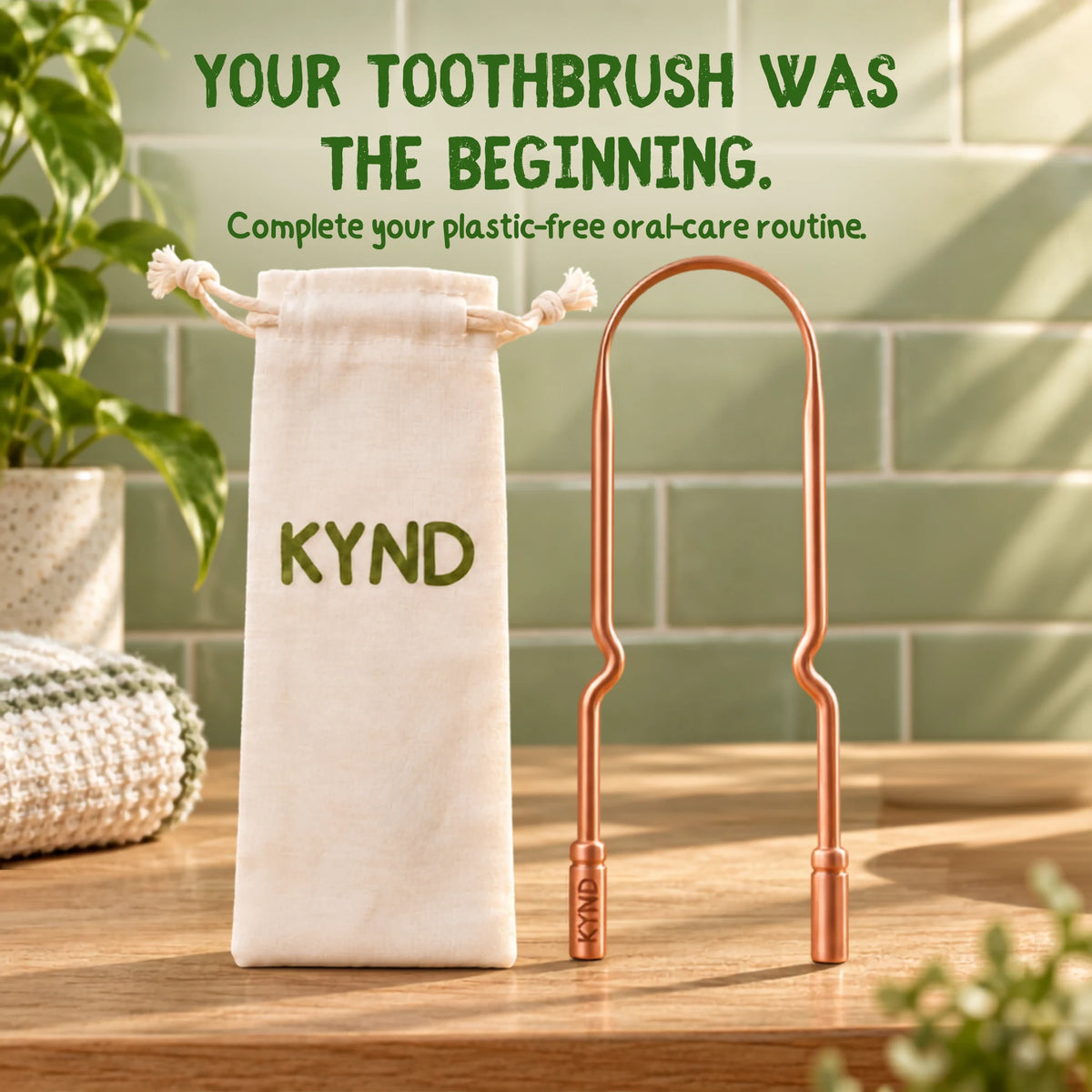 A copper toothbrush holder with a wavy handle stands next to a cream drawstring pouch printed with the brand name KYND, set on a wooden table against a green tiled wall background. Text at the top reads, 'YOUR TOOTHBRUSH WAS THE BEGINNING. COMPLETE YOUR PLASTIC-FREE ORAL-CARE ROUTINE